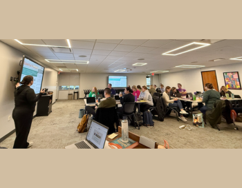 Presenter at the front of a conference room pointing to a projection screen, with attendees seated at tables looking towards the screen. The room is well-lit and features colorful wall art.