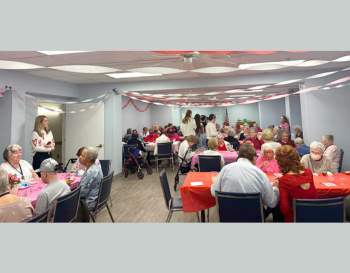 Group of people at a Valentine's Day party in a decorated room with pink and red streamers, some seated at tables covered in red cloths and others standing, socializing.