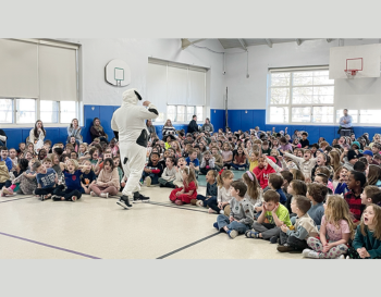 An individual dressed as a dog engages with school children seated on a gym floor during an event.