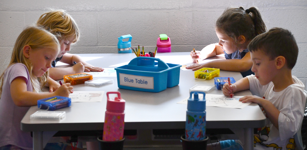 Four children are sitting around a table, engaging in drawing activities. A blue bin labeled "Blue Table" with art supplies and water bottles are also on the table. The setting appears to be an indoor classroom.