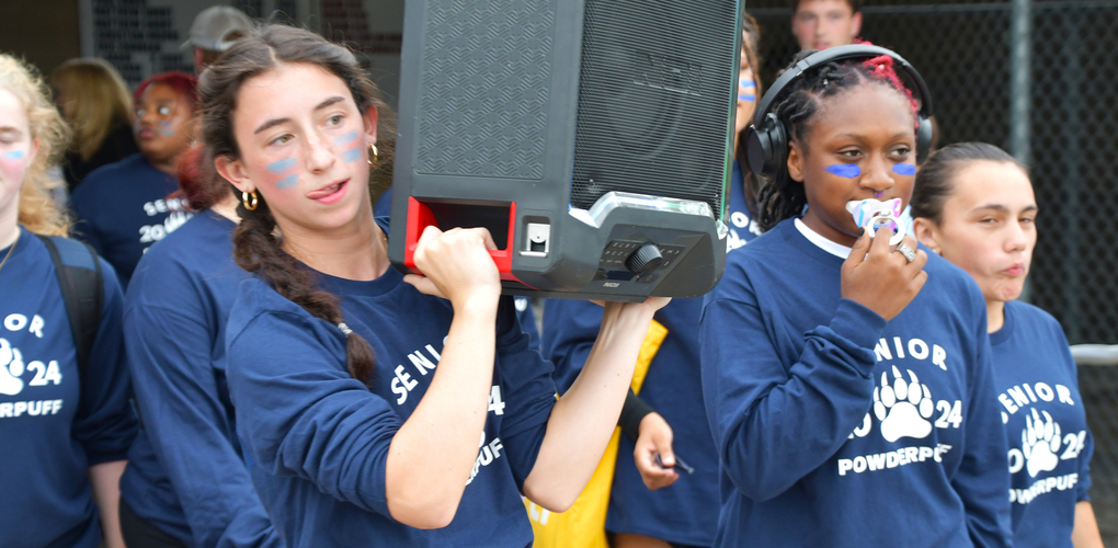 Person carrying a speaker on their shoulder in a crowd of people wearing 'Senior Powderpuff' t-shirts.