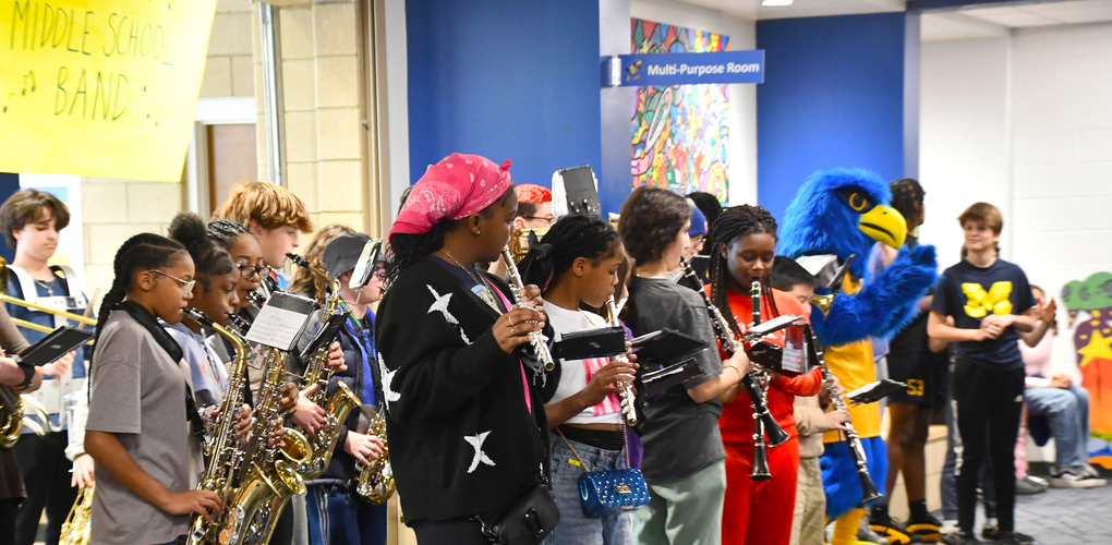Students playing various musical instruments in a school hallway with 'Middle School Band' and 'Multi-Purpose Room' signs visible in the background.