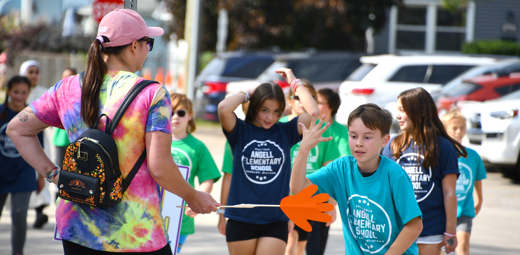 Participants at a fun run event, with one person in a tie-dye shirt facing a group of children wearing 'Angell Elementary School' t-shirts, in a suburban setting.