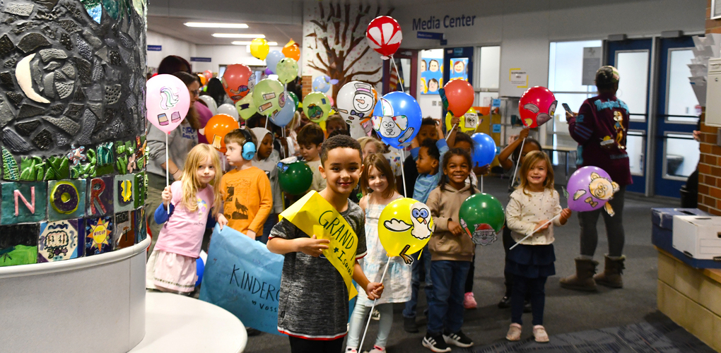 Children holding balloons decorated with various designs, standing in a school media center.