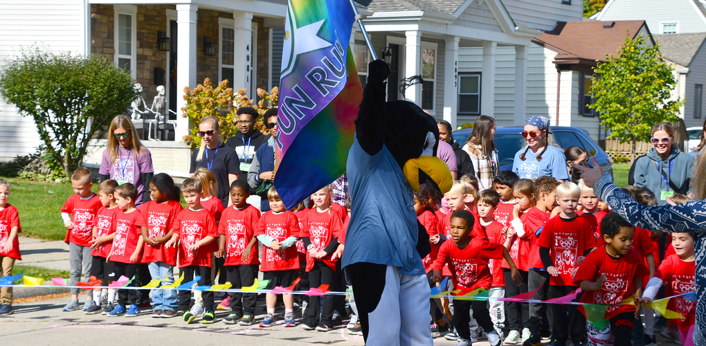 Group of children wearing red shirts participating in a Fun Run event, starting to run behind a mascot holding a rainbow flag, in a residential street setting.