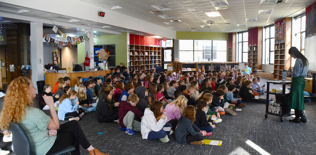 A group of students sit on the floor of a library, listening attentively to a presenter who is standing. The library features bookshelves and bright, colorful decorations.