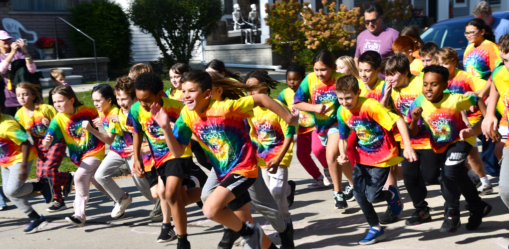 A group of children wearing colorful tie-dye T-shirts with "Fun Run" logos participate enthusiastically in a running event, with a sunlit street and spectators in the background.