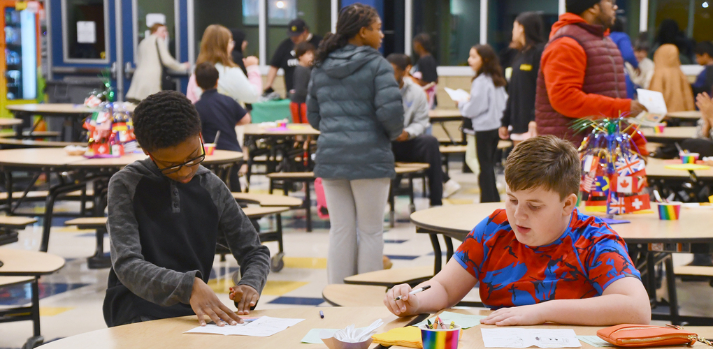 Students engaged in activities at a school cafeteria with others walking and interacting in the background.