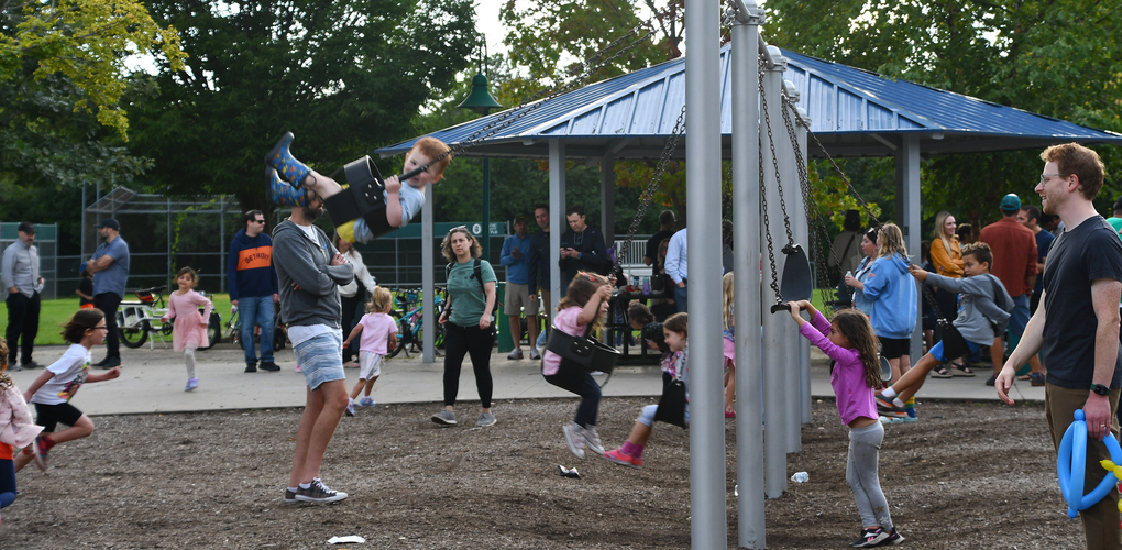 Adults playing with children by pushing their swings into the air on a busy playground, with other children and adults around, some interacting and others moving around.