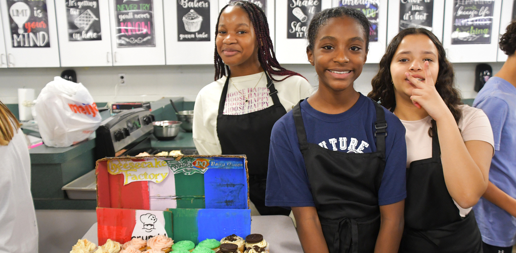 3 students posing in a classroom kitchen and smiling by their decorated cupcakes during a cupcake wars competition