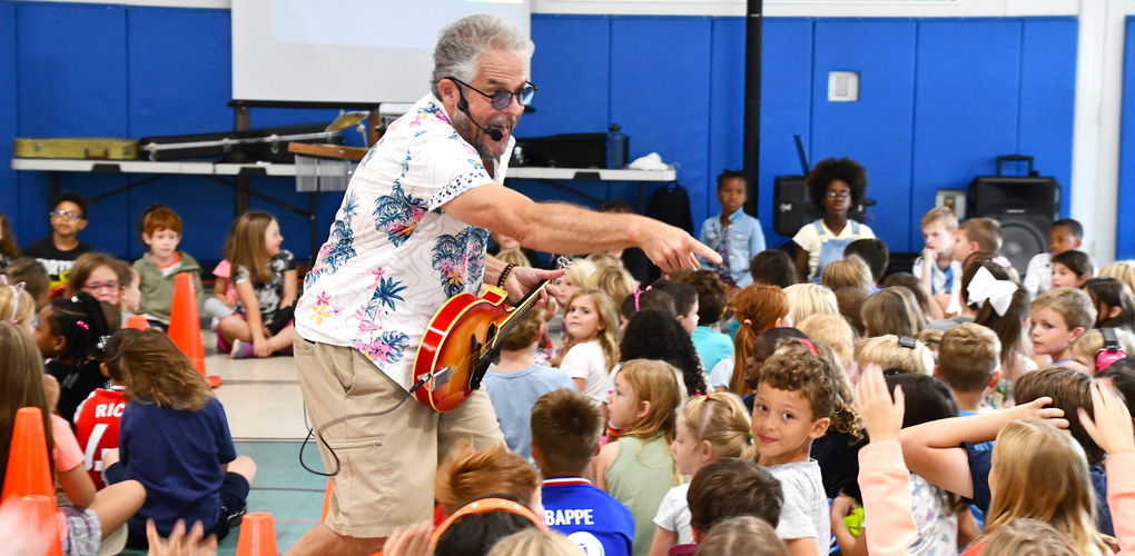 Musician performing with a guitar for an enthusiastic group of elementary school children in a school auditorium.