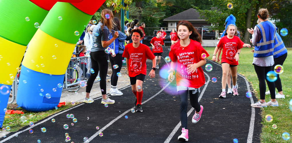 Participants in red shirts running through a finish line under a colorful arch with bubbles floating around at a race event.