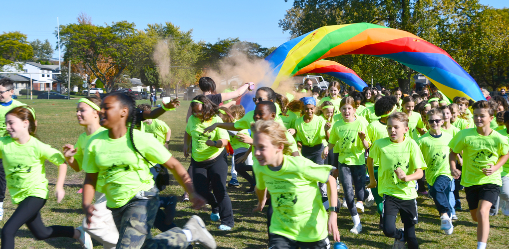 Children in green shirts participating in a playful outdoor event, running under a large, colorful arch on a sunny day.